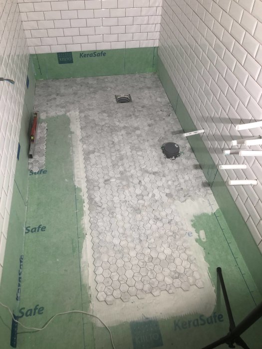 Pågående installation av hexagonala klinkerplattor på badrumsgolv med torrlagda bitar runt golvbrunn.