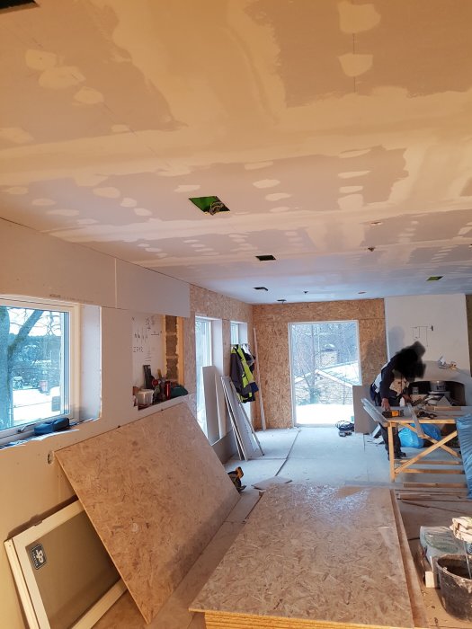 Renoveringsarbete i ett rum med spacklade väggar och tak, byggmaterial och en arbetare.
