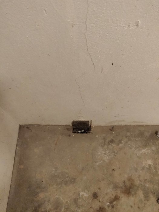 Gammal, liten fyrkantig lucka i ett källargolv, nära en vägg med spricka och slitet betonggolv.