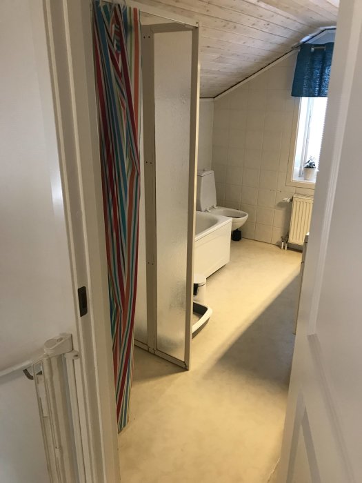 Badrum före renovering med toalett och dusch, vit kakel, trädetaljer och färgglad duschdraperi.