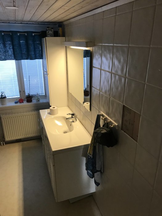 Badrum före renovering med handfat och spegel, kaklade väggar och fönster med mörkblå gardiner.