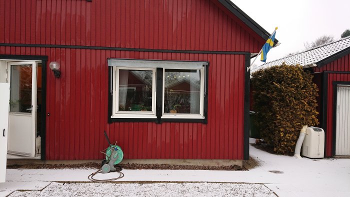 Rödfärgad träfasad på hus med fönster, vit dörr och en svensk flagga, lätt snötäckt mark.