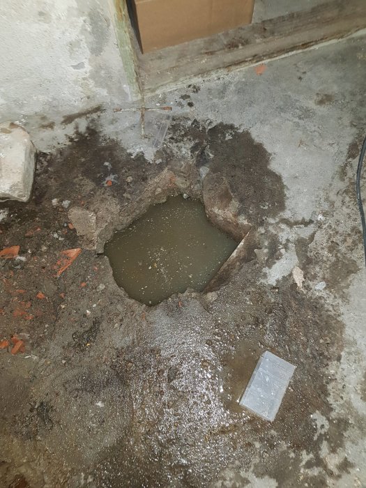 Ett hål i ett betonggolv fyllt med vatten där ett avloppsrör har gått sönder, omgivet av grus och smuts.