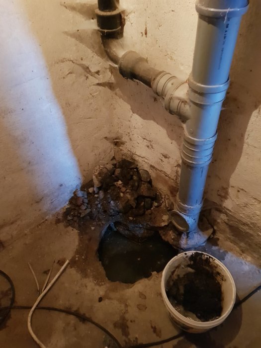 Hål i betonggolv med ett trasigt avloppsrör, omgivet av grus och en hink med avlagringar.