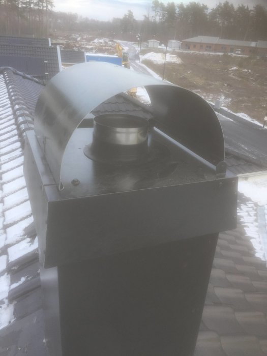 Takutsikt med en nyinstallerad kamin och skorsten, omgiven av en halv snötäckt tak och skogsbakgrund.