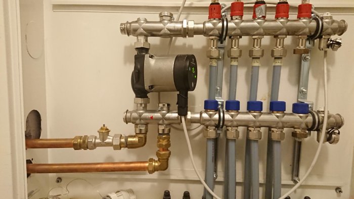 Golvvärmeinstallation med en Grundfos Alpha2 cirkulationspump, kopparledningar, och en golvärmefördelare med flera blå ventiler.