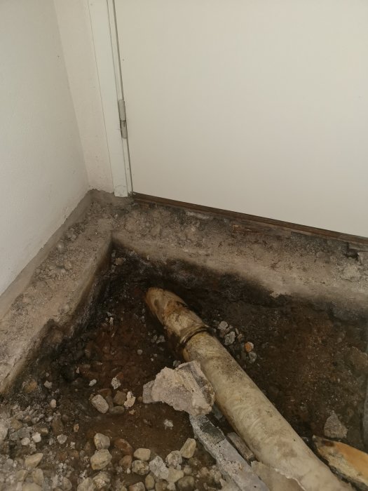 Öppnat golv i ett hörn av en källare som visar delvis exponerade avloppsrör, inklusive ett gammalt vitt PVC-rör.