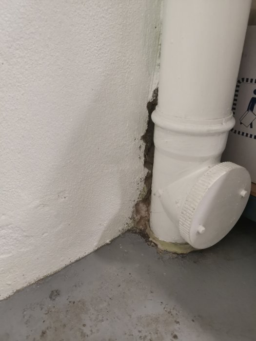 Vitt PVC-avloppsrör som leder in i ett betonggolv med synlig anslutning och borrhål i källare.