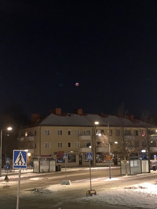 Tidig morgonfoto av en gata med snö, byggnader och trafikskyltar under en röd månförmörkelse på himlen.