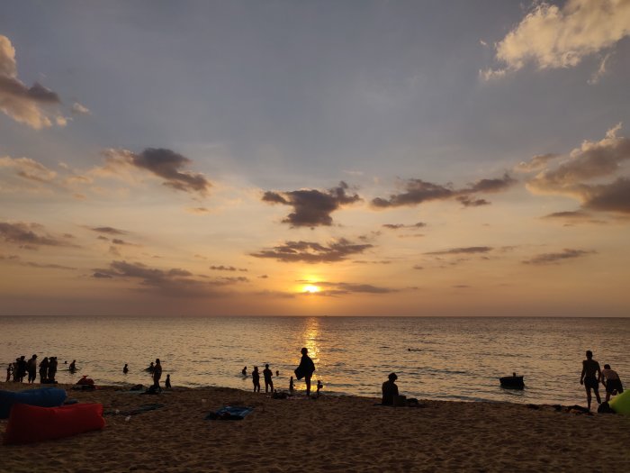 Solnedgång på en strand med människor som badar och umgås, med moln på himlen.