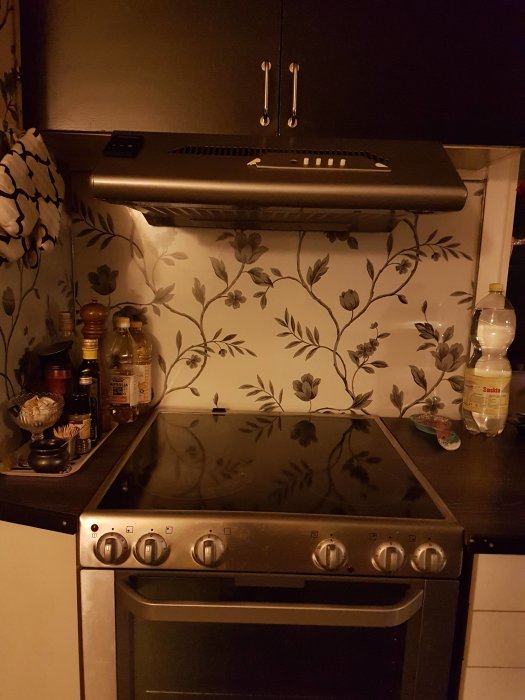 Kökshörna med spishäll, fläkt och små köksredskap mot en blommig vägg.