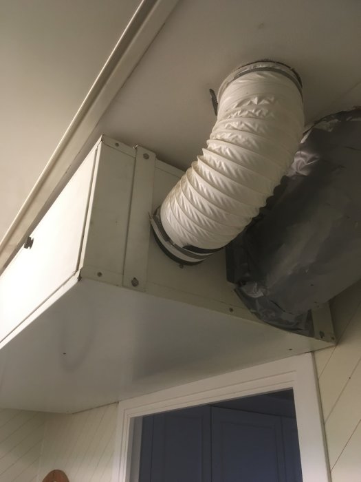 Vit ventilationskanal som går genom en vit lucka i taket över en blå dörr.