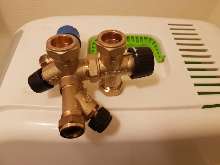 Kopparfärgad blandningsventil med två svarta rattar på en vit yta, nära grönt föremål, med R15 vattenuttag synliga.