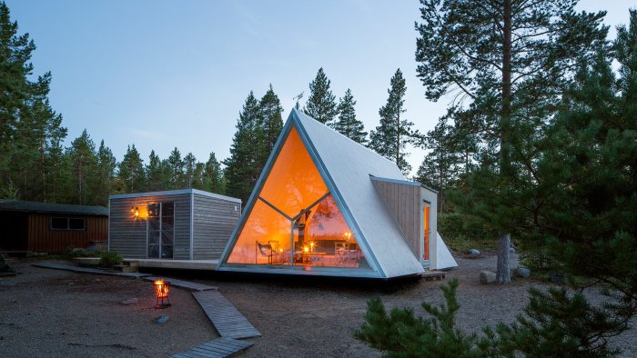 A-frame hus med belyst interiör i skogsmiljö i skymningen, kännetecknat av en spetsig takkonstruktion.