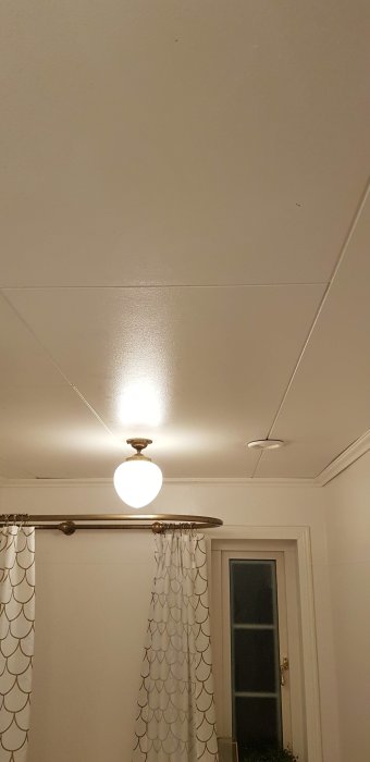 Vitt slätt badrumstak med taklampa och draperistång ovanför vit dörr.