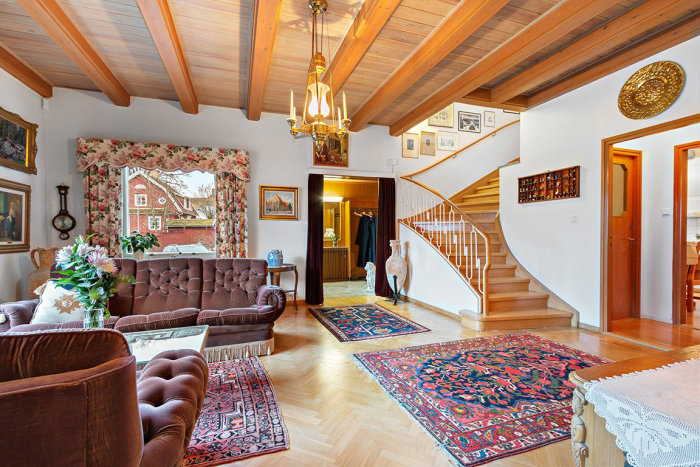 Välbevarat vardagsrum i villa med träbjälkar, trappa, mönstrade mattor och klassisk inredning.