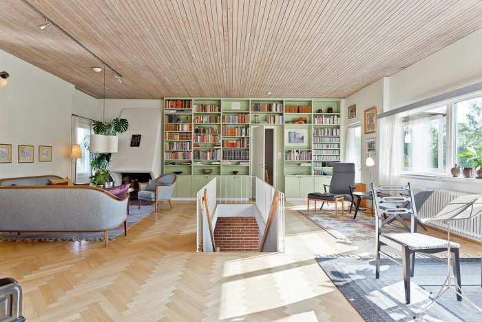 Retro vardagsrum med trägolv, paneltak, gröna bokhyllor längs väggen, moderna möbler och stora fönster.