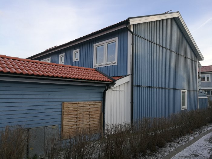 Exteriör av ett blått radhus med olikformade tak och en liten, plåtbeklädd förrådsbyggnad till vänster.