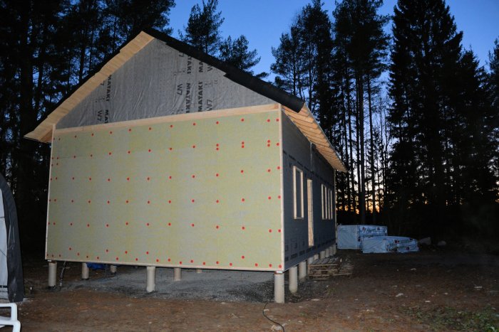 Hus under konstruktion med klimatskivor och stående läkt på gavelsidan vid skymning.