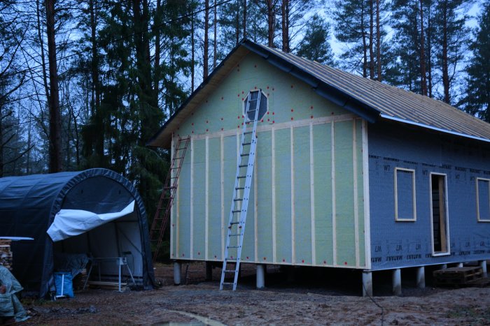 Hus under renovering med klimatskivor och påbörjad stående panel på gavelsidan, stege lutad mot väggen.