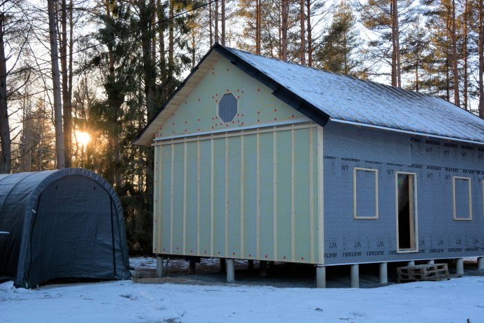 Hus under uppförande med halvklar gul panel och klimatskivor i ett vinterlandskap vid solnedgång.
