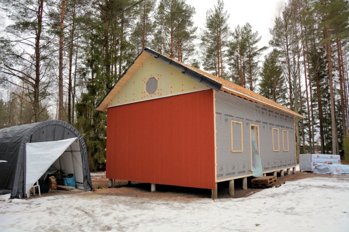 Hus under renovering med ena gavelsidan täckt av röd panel, andra sidan med isolering och läkt.