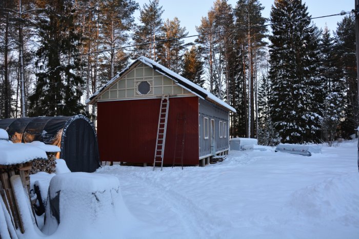 Hus under renovering med nästan färdig exteriör panel, stegar mot vägg, omgivet av snötäckt landskap.