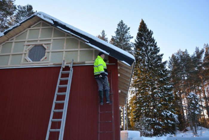 En person i arbetskläder på en stege vid en husgavel som är delvis täckt av snö, med synlig isolering och obehandlat virke.