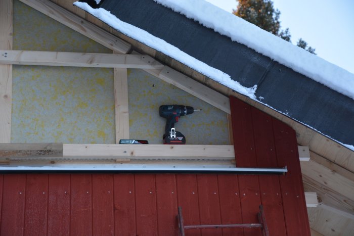En del av en husfasad med röd panel och synlig isolering ovanför, med ett batteridrivet borrskruvdragare på en trälist under ett snötäckt tak.