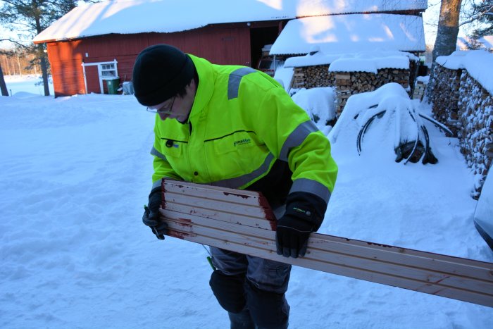 Person i arbetskläder inspekterar målad träpanel utomhus i snöig miljö.