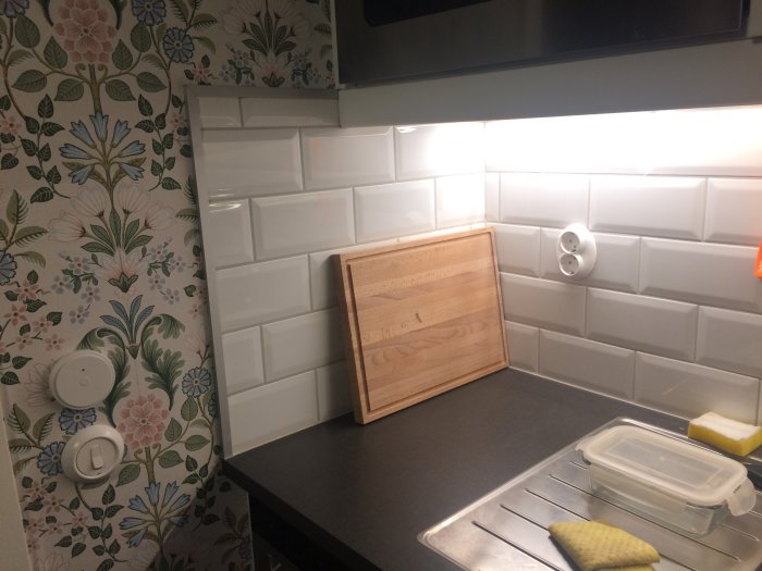 Kökshörn med vit metrokakel, trälist och mönstrad tapet ovanför bänkskiva med diskbänkstillbehör.