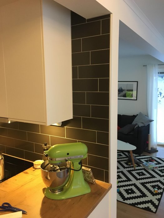 Köksmiljö med kakel i mörkt utförande som fortsätter upp till taket, med en grön köksassistent på bänkskivan.