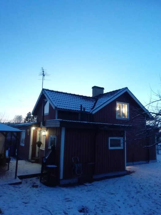 Ett tvåvånings trähus i skymningen med snö på marken och en antenn på taket till den äldre utbyggnaden till vänster.