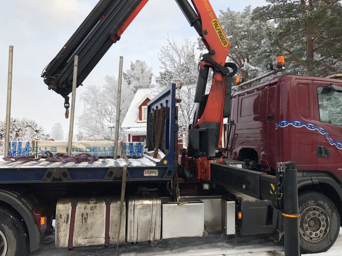 Lastbil med kran lastar av byggskivor i snöigt landskap, indikerar leverans av material för takrenovering.