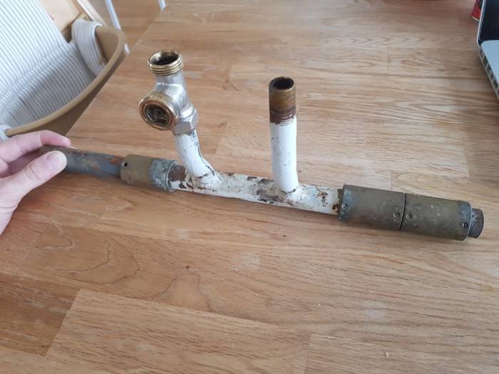 Hand som håller en gammal avloppsrörinstallation med korrosion och två anslutningsmuttrar mot ett trägolv.