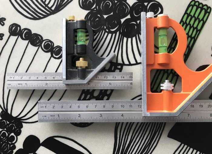 Två snickarvinklar, en svart och en orange, med inbyggda vattenpass och linjal med cm och tum.