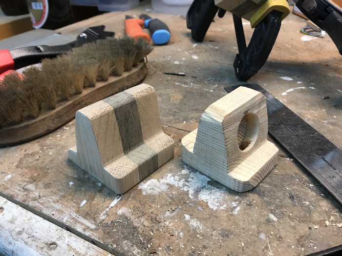 Hemmagjord Apple Watch-laddarhållare i ek vid sidan om ett furumodell, på ett stökigt arbetsbord.