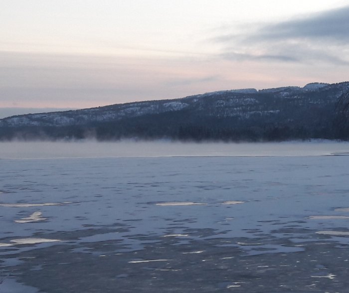 Isfrusen havsyta med sjörök och skogklädda berg i bakgrunden vid kyligt vinterväder.