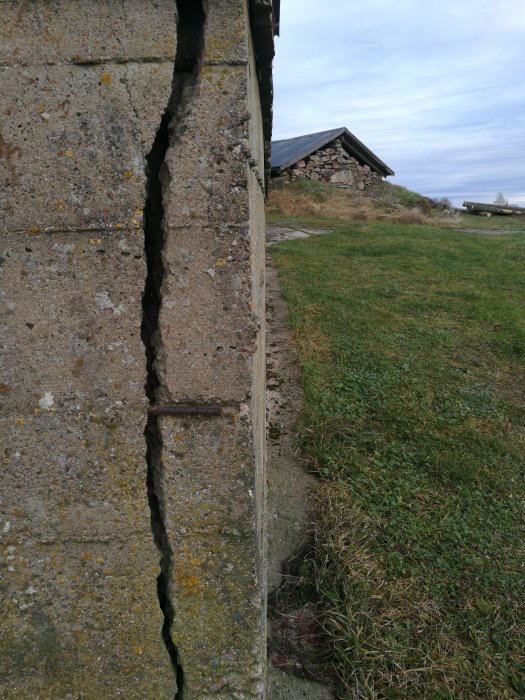 Stor spricka i en betongmur utomhus med armeringsjärn synligt, samt en del av en byggnad i bakgrunden.