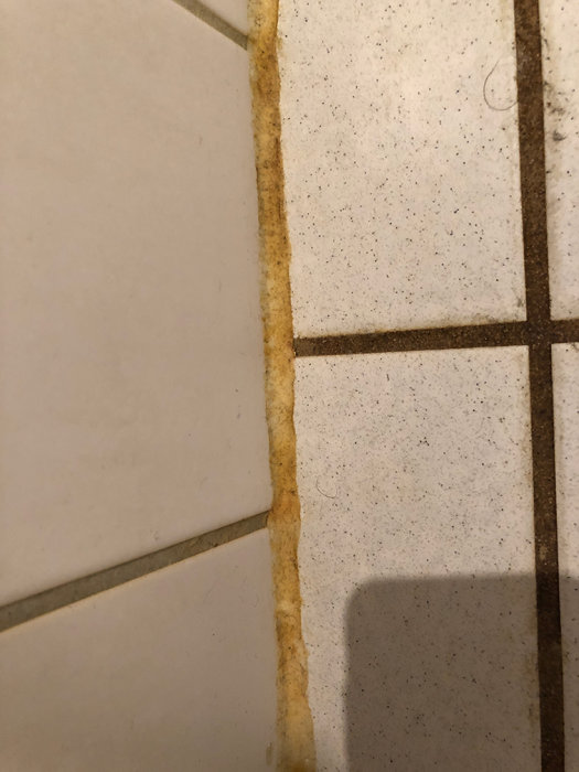 Gult missfärgade fogar mellan vita kakelplattor på ett badrumsgolv.