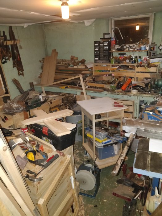 Rörig verkstad med utspridda verktyg, trämaterial och byggutrustning, osäkert byggnadsframsteg.