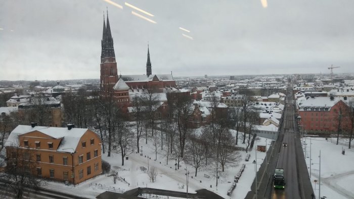 Utsikt över en snötäckt stad med en kyrkspira, träd och byggnader, tagen från ett fönster.