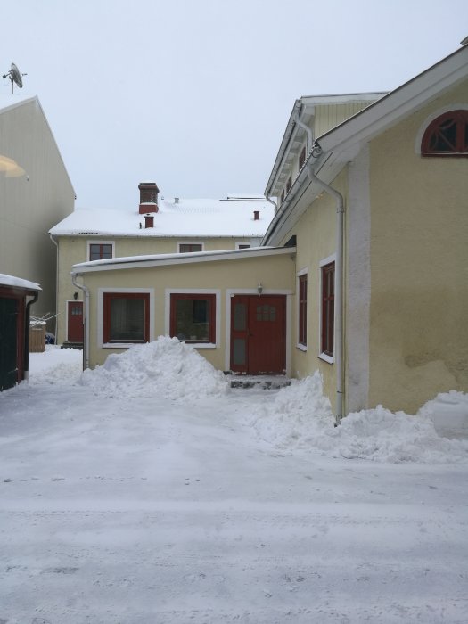 Gult hus med röda detaljer täckt av snö, snödrivor framför huset på en plogad väg.