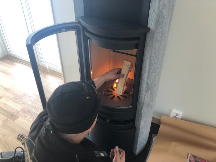 Person lägger in vedträ i en modern kamin med synlig flamma och stenbeklädnad.