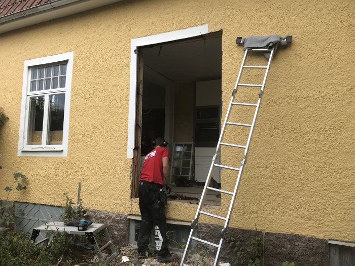 Person monterar altandörr i en gulputsad fasad med stående plankkonstruktion, intill ettfönster från 1928.