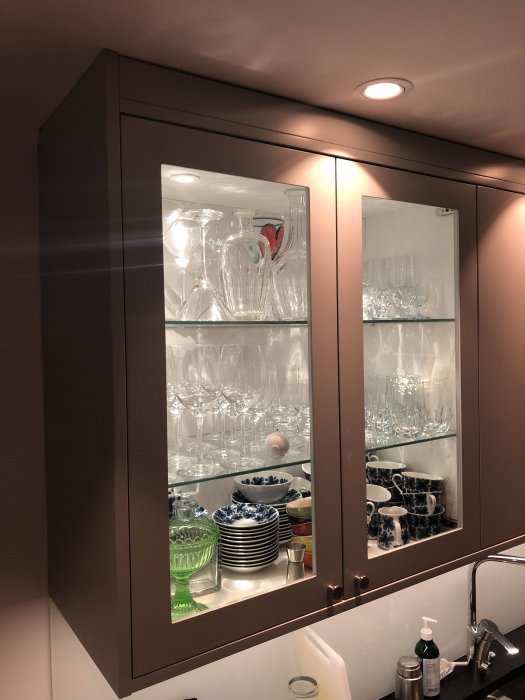 Väggmonterat vitrinskåp med glasdörrar, fyllt med glas och porslin, ovanför köksbänk.