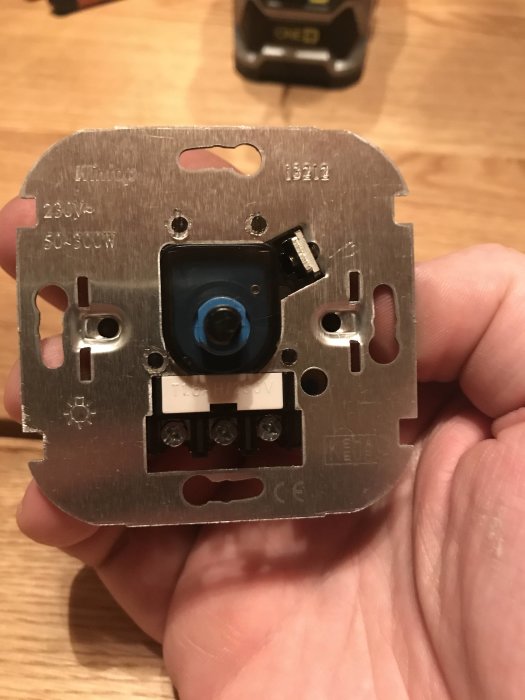 Hand som håller en avmonterad dimmerbrytare med etiketter och anslutningsterminaler synliga, redo för elinstallation.