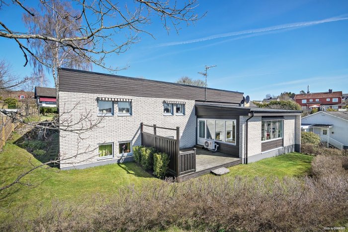 Vitputsat hus med svarta tak och träpanel, gräsmatta och buskar i ett solbelyst bostadsområde.