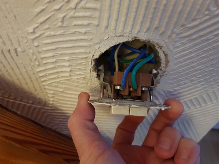 Hand håller en borttagen eluttagsfront vid ett öppet uttag med synliga kablar i en vägg med spackelstruktur.