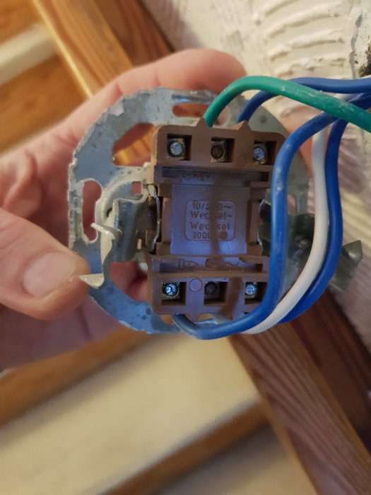 Hand håller en elektrisk väggkontakt med synliga blå och gröna ledningar och en metallmonteringsplatta.